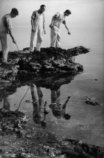 Long shot of three men, wearing overalls, standing on a rocky outcrop surrounded by water. At the Beach, Circa 1955