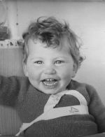A close up shot of a young child smiling at the camera. The child is wearing a jumper with white tape over the jumper. The number 4 is stamped on the tape. Portrait of a Child, Circa 1955