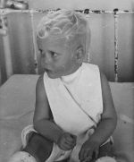 A close up shot of a young child sitting in a bed with casts on their legs, up to their knees. Portrait of a Child, Circa 1955