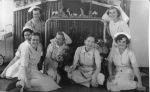 A medium shot of a seven nurses. The five nurses in the foreground are sitting on the floor, and the two nurses in the background on the left and right sides are kneeling. Nurses Posing in Front of the Nativity Scene, Circa 1955