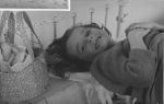 A close up shot of a young child lying on a bed, smiling at the camera. The child has their hand crossed on their chest and there is a basket on a cupboard on the left hand side of the photograph. In Bed, Circa 1955