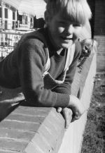 Close up shot of two children smiling for the camera. Both are leaning on a brick wall with part of a building in the background and a large shade umbrella. On the Balcony, Circa 1955