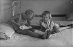 A long shot of two children on a bed. One is sitting on the bed, and cutting paper with scissors. The other child is half on, half off the bed, smiling for the camera with their face propped up with one hand. Playing in Bed, Circa 1955