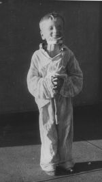 A long shot of a child smiling for the camera. The child is wearing baggy pyjamas and has a brace around their neck that is propping up their chin. Pyjama Time, Circa 1955