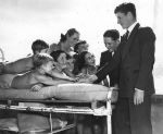 Medium shot of seven children outside, three laying on a bed, four standing beside the bed all arms stretched out clasping the hand of a person standing at head of bed, another person is standing alongside the bed. Visitors to the Hospital, Circa 1955