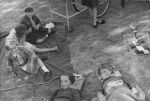 Medium shot of five children outside, three are seated two are in girl guide uniforms, the remaining two are laying supine on stretchers, a flower bed with toadstool garden ornament is visible amongst the flowers left of the image. Relaxing on the Lawn, Circa 1955