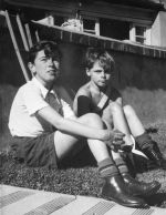 Medium shot of two children posing for the camera. They are sitting on grass, with a brick wall in the background. The child in the foreground, has their arms wrapped around their knees and is wearing a collared shirt, shorts, socks and boots. Patients on the Lawn, Circa 1955