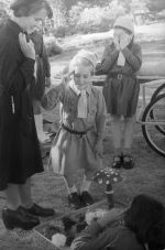 Long shot of a person in a girl guide uniform, two children are standing in girl guide uniforms one child is saluting, the other child face is obscured by their hands, another child is laying supine saluting, a flower bed with a toadstool garden ornament amongst the flowers below centre of the image. Brownies, Circa 1955