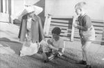 Long shot of a nurse with cape on kneeling down outside on a grassed area, alongside one child is seated looking at a book wearing a caliper, another child is standing nearby. Reading Outside, Circa 1955