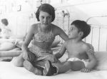 Medium shot of two children on a bed, one child is kneeling on the bed with an arm around the other seated child's back and arm. In the left background another child is seated on a bed. Ward Scene, Circa 1955