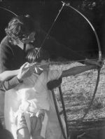 Medium shot of an adult outside aiding a child to play archery. The child has a brace secured by straps from shoulder to hip and waist, the other child's arm is supported by a crutch. Archery Lessons, Circa 1955
