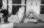 Medium shot of a child outside laying supine on a bed reading a book with one leg raised at the knee. Reading in Bed, Circa 1955