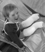 Medium shot of a child outside seated on a blanket, braces around neck and chest both legs have plaster casts from above knee to toes posing for the camera. Portrait of a Child, Circa 1955