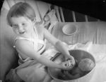 Medium shot of a child with braces from shoulder to waist seated on a bed washing a doll in a large bowl posing for the camera. Playing in Bed, Circa 1955