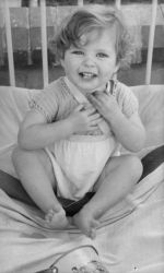 Medium shot of an infant seated on a bed arms on chest posing for the camera. Portrait of a Child, Circa 1955