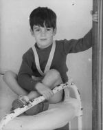 Medium shot of a child seated on a bed legs crossed one hand on the bed railing the other hand holding another structure for support posing for the camera. Portrait of a Child, Circa 1955