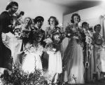 Long shot of a wedding party scene. Appears to be up on stage. The bride is carrying a bouquet of flowers, and a horseshoe on a ribbon. The women and children in the foreground are also carrying bouquets of flowers. There is one male in the photograph, in the background, who is wearing a uniform and cap. Pantomime at the Hospital, Circa 1955