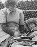 Mid shot of a nurse outside seated on a bed reading a book to a child laying on a bed. In the foreground is an abacus wooden toy. Nurse Reading to a Child, Circa 1955