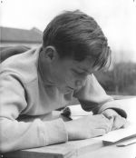 Medium shot of a child outside laying down doing homework with a pencil and paper. Reading, Circa 1955
