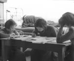 Medium shot of three children seated at a desk writing on paper a bed is in the background. Reading and Writing, Circa 1955