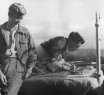 Medium shot of two adults outside one is seated on edge of bed, the other is laying on the bed writing on paper. Reading and Writing, Circa 1955