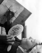 Medium shot of a child laying supine on a bed painting, holding a mirror with an easel positioned above the bed, a paint palette in the foreground. Painting, Circa 1955