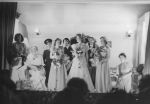 Long shot of a wedding party scene. Appears to be up on stage as the backs of people's heads are in the foreground. There are women and children carrying bouquets of flowers. There is one male in the photograph, in the background, who is wearing a uniform and cap. Pantomime at the Hospital, Circa 1955