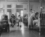 Long shot of a children's ward. To the left is a young patient sitting in a wheelchair reading a book, in the centre are three young patients sitting at a table drawing and to the right are beds with nurses attending to the patients. Ward Scene, Circa 1955