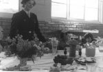 Medium shot of a woman wearing a uniform standing behind a table set with party food and flowers. There are three children sitting beside her wearing party hats. Birthday Celebrations at the Hospital, Circa 1955