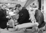 Medium shot taken outside of a young patient sitting on a bed wearing a shiny fireman's helmet. There are two men in uniform helping as Father Christmas gives a present. Fire Brigade Visits, Circa 1955