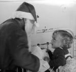 Close shot of the back of Father Christmas handing a present to a young patient lying face down on a metal framed bed. Father Christmas at the Hospital, Circa 1955