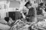 Medium shot of a young patient lying in a bed watching a woman standing beside writing in a book. There are other patients and adults in the background. Jenny Howard Visits, Circa 1955