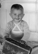 Close shot taken from the front of a young child smiling at the camera sitting on a bed holding a box on the lap. Christmas Time, Circa 1955