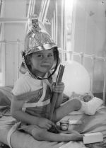 Close shot of a young child sitting on a metal framed bed wearing a shiny fireman's helmet. There is a cup of Peter's ice-cream sitting between the legs and one ankles is in plaster. Christmas Time, Circa 1955