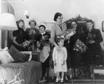 Long shot of a group of women holding bouquets standing in a lounge room setting. One woman has one arm resting on a young child. Pantomime at the Hospital, Circa 1955