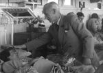 Medium shot of a patient lying in a bed with a man in a suit standing beside reaching out a hand to the pillow. There are other children in beds in the background. Uncle Bobs Club Visiting, Circa 1955