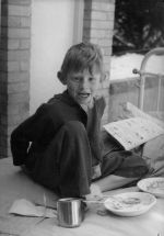 Medium shot of a young patient sitting outside on a metal framed bed. There is a comic on the bed as well as two empty bowls with spoons and a metal cup. Dinner in Bed, Circa 1955