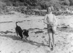 Long shot of a child dressed in shorts on the beach holding a plastic bucket. There is also a dog playing behind. At the Beach, Circa 1955