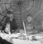 Medium shot of two young children sitting under a beach umbrella enjoying bowls of food. Under the Sun Umbrellas, Circa 1955