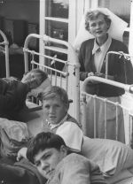 Medium shot of three young patients lying on metal framed beds with a nurse standing behind. Patients and a Nurse, Circa 1955