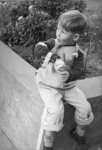 Medium shot of a young child sitting on the concrete edge of a garden bed. The left arm is in a frame. Portrait of a Child, Circa 1955