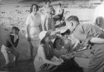 Long shot of a group paddling at the beach. In the foreground are some children wearing sun hats and laughing with a man in uniform while two women watch. In the background a child sits in a wheelchair on the sand. Greeting Visitors, Circa 1955