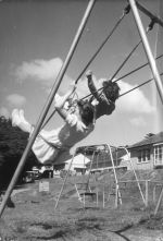 Long shot taken outside of two children flying high on a swing set. Playing on the Swings, Circa 1955