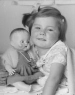 Close shot of a young child looking at the camera and holding a doll. The doll has a knitted dress and cardigan and wearing pearls. Portrait of a Child, Circa 1955