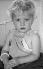 Close shot of a young child sitting on a metal framed bed looking unsure at the camera. There is a beaded bracelet on the right wrist. Portrait of a Child, Circa 1955