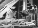 Medium shot taken outside of a patient lying face down on a metal framed bed. A nurse is attending to their back while another young person leans through the bedhead, reading a book. Nurse and Patients, Circa 1955