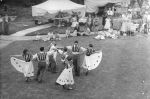 Long shot taken outside from above of four couples dressed in costume dancing a reel. There are people sitting on the lawn or standing in the background watching. At the Races, Circa 1955