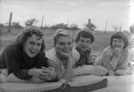 Medium shot taken outside of four young patients lying on cushions on the ground smiling at the camera. At the Races, Circa 1955