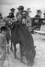 Long shot of a man helping a young child get off a horse. The child has a leg in callipers. There is another child dressed in riding gear standing and holding reins. At the Races, Circa 1955