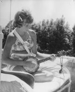Medium shot of a young woman dressed in a two piece bathing suit sitting on a bed outside playing a banjo. Playing Musical Instruments, Circa 1955